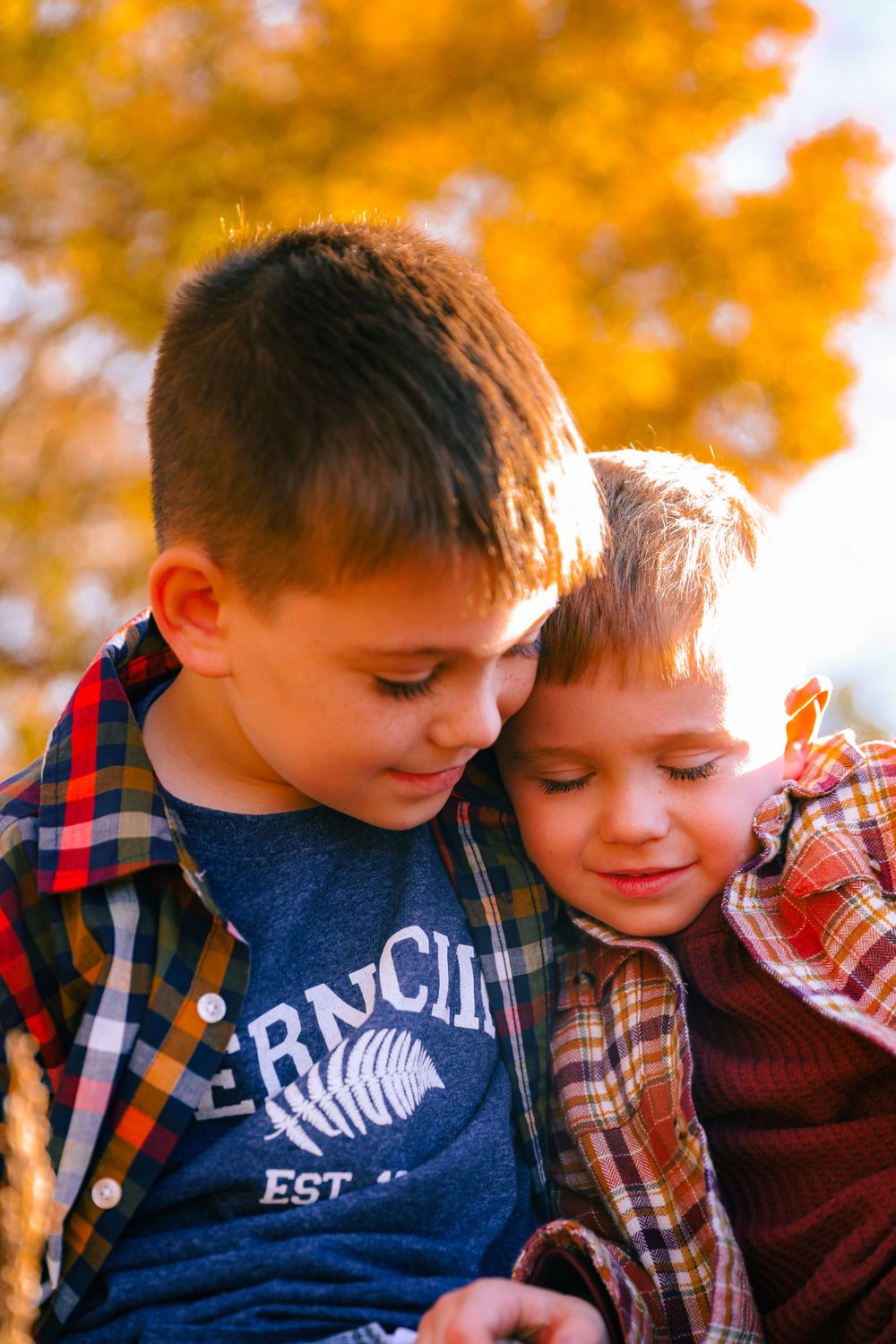 Two young siblings in plaid shirts share a sweet moment against a backdrop of autumn foliage.