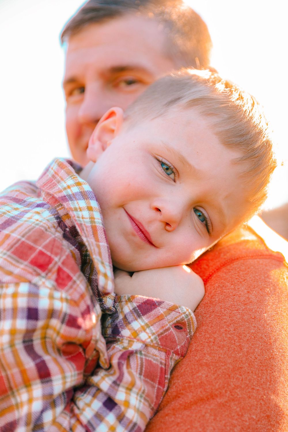 Child in plaid shirt rests head on shoulder during golden hour sunset.