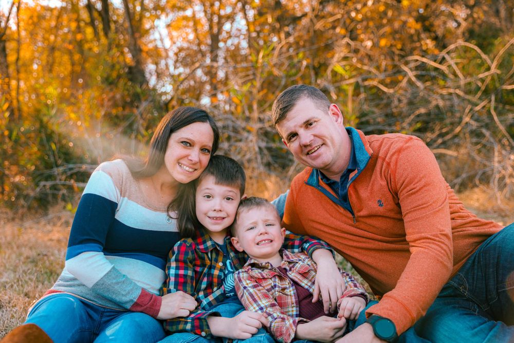 Family of four poses together on ground with autumn foliage in background.