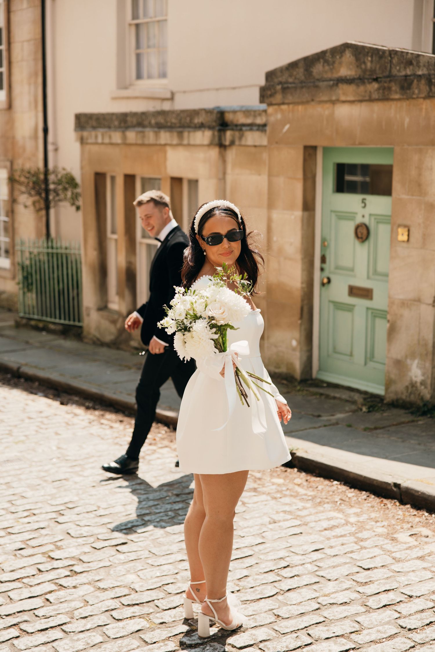 Stylish bride in short white dress holding bouquet on cobblestone street.