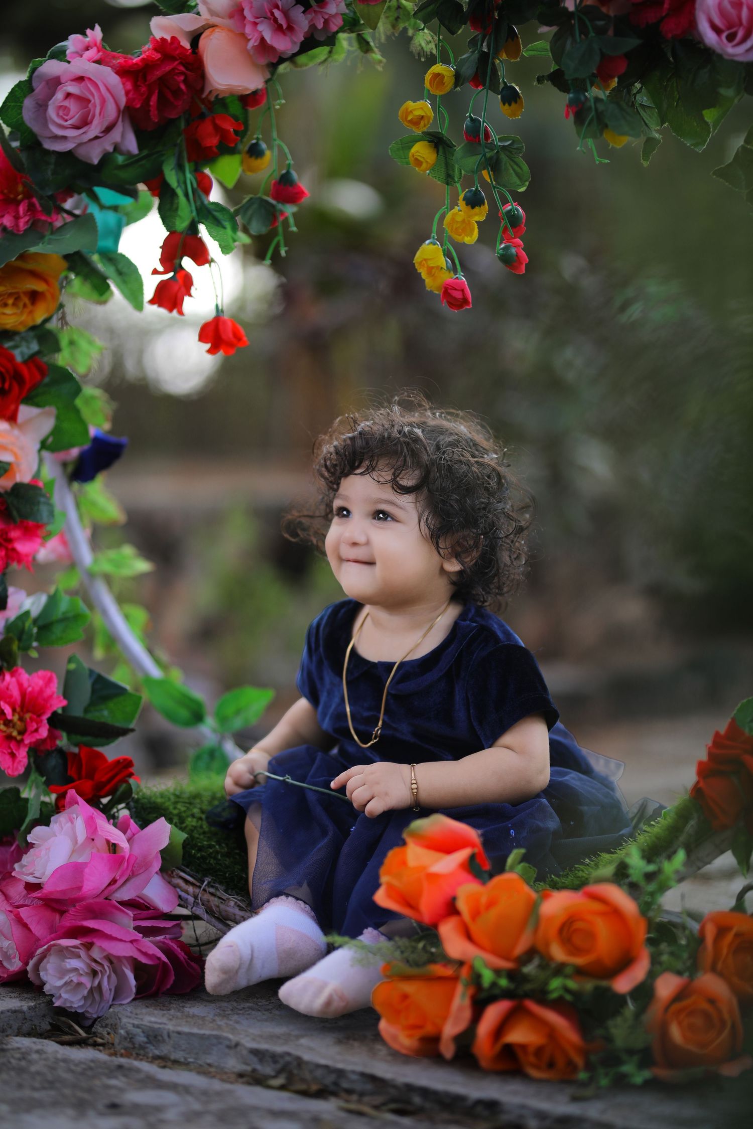 Curly-haired toddler in navy dress sitting among vibrant roses and floral decorations in a garden setting.