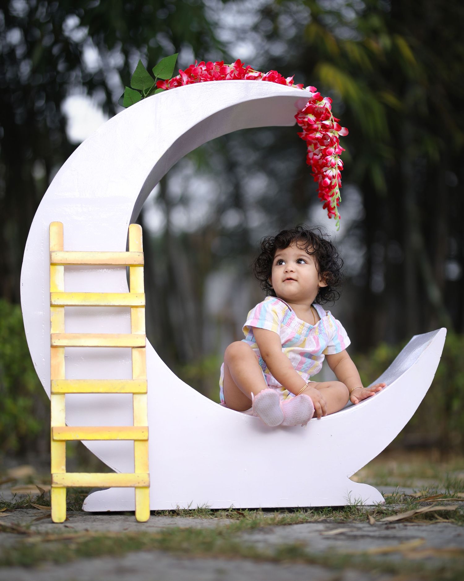 Small child sitting on decorative white crescent moon prop with yellow ladder and red floral accents.