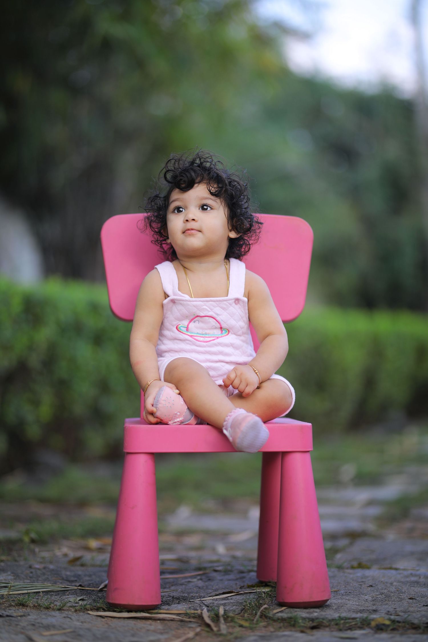 Small child seated on bright pink chair outdoors surrounded by greenery.