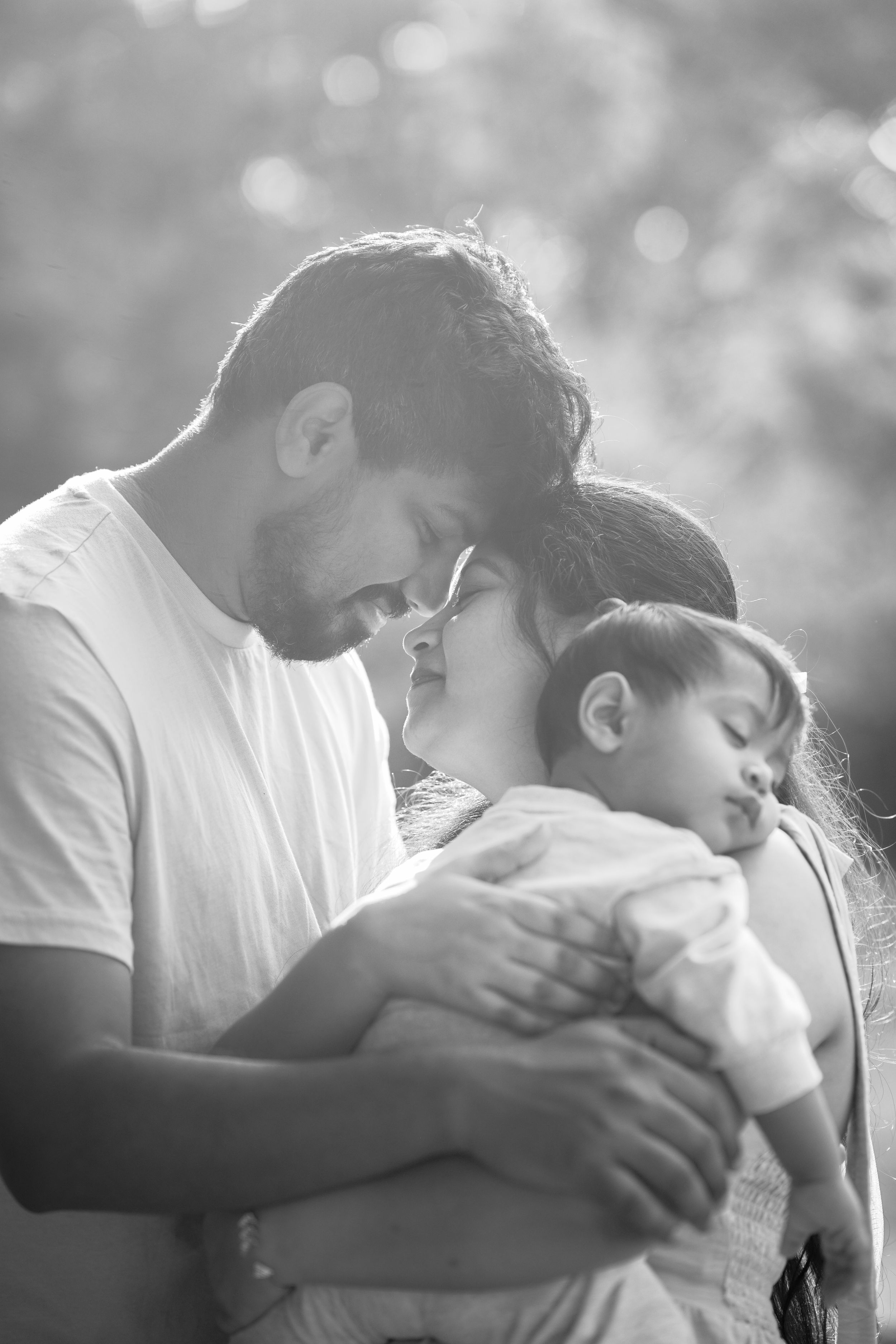 A tender family moment captured in black and white showing parents embracing their sleeping child in soft natural light.