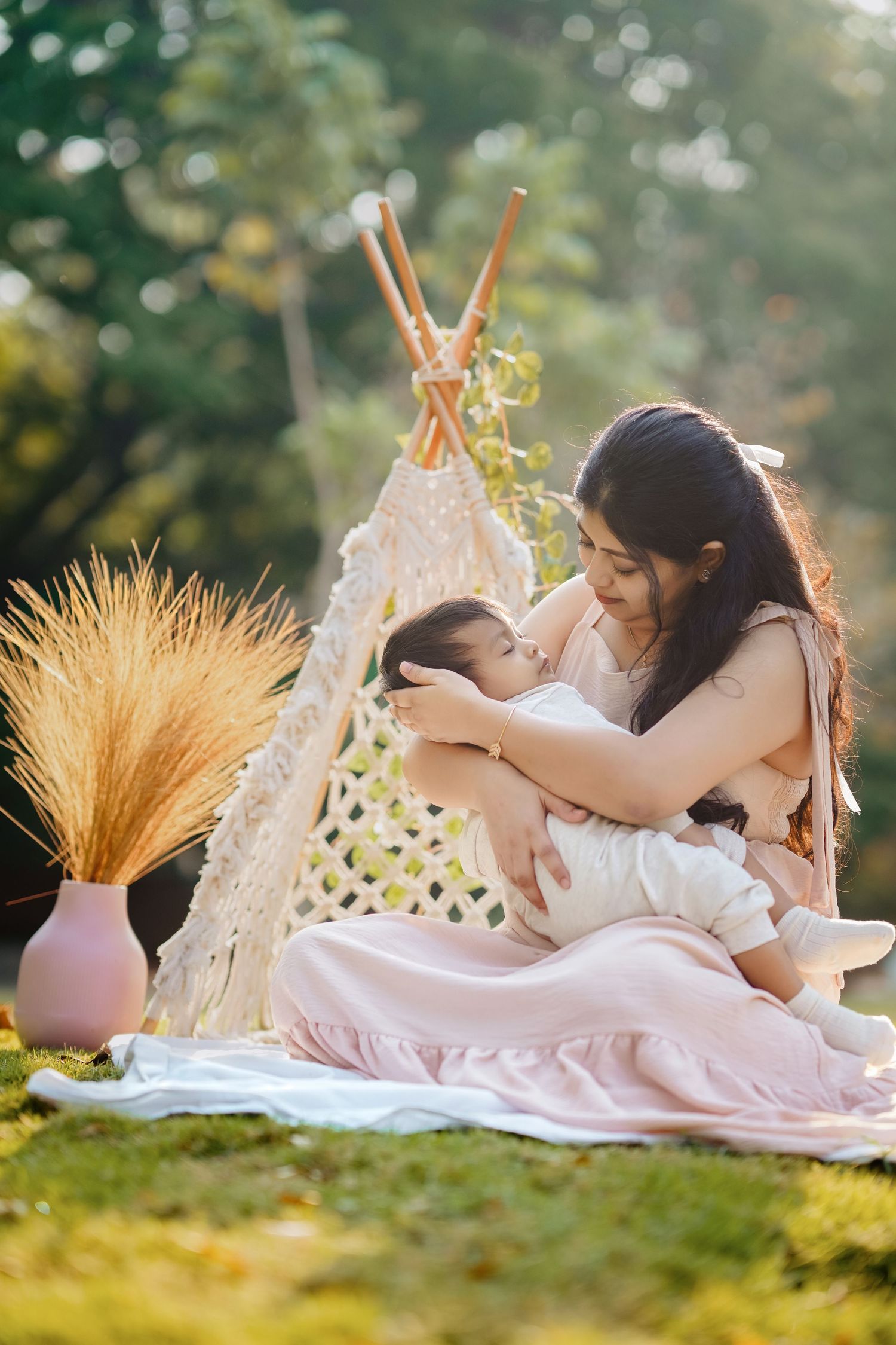 A tender outdoor picnic scene with caregiver and child on blanket next to decorative teepee and pampas grass at sunset.