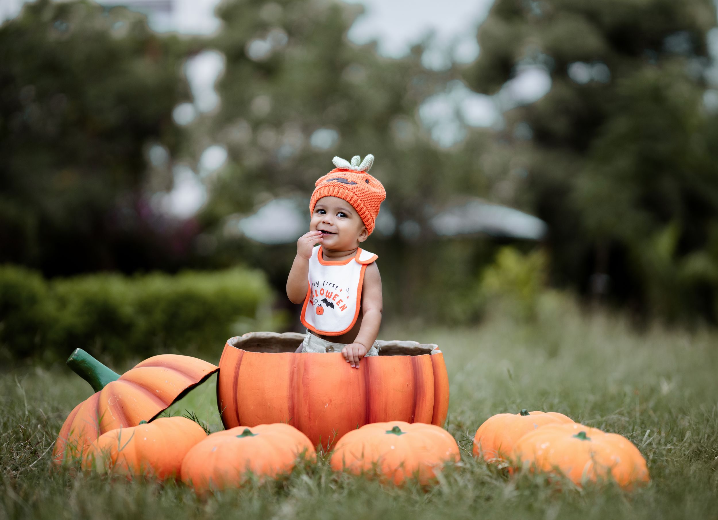 Baby in pumpkin hat sitting inside a large pumpkin prop surrounded by orange pumpkins on green grass.