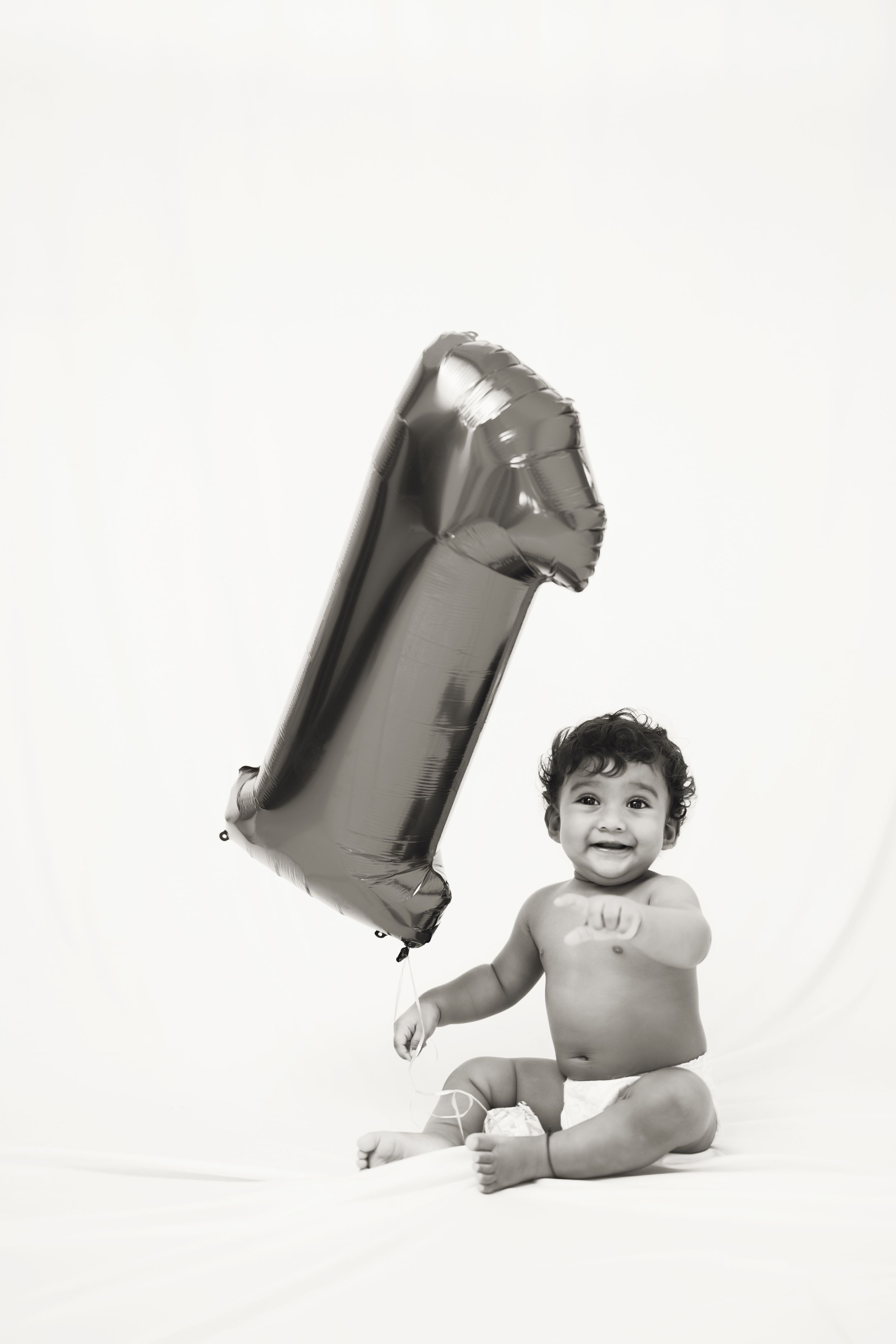 A smiling baby sits beside a number 1 balloon, celebrating their first birthday in a black and white photo.