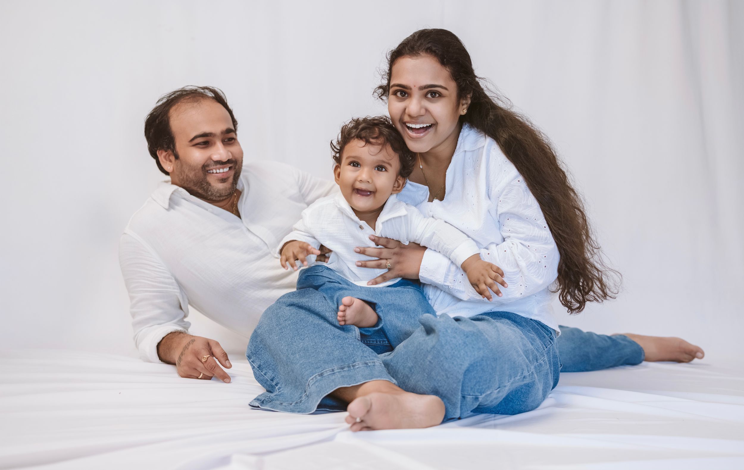 Happy Indian family with parents in white shirts and jeans sitting on white backdrop with smiling baby.