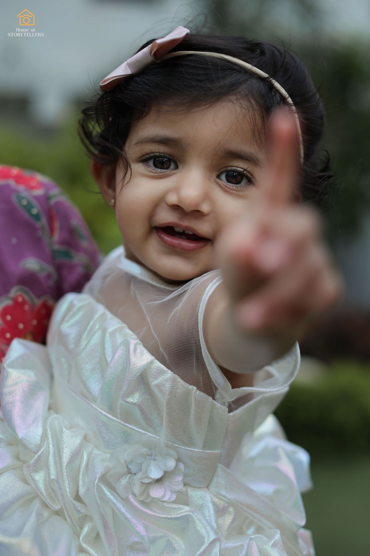 Small child in white dress pointing playfully at the camera.