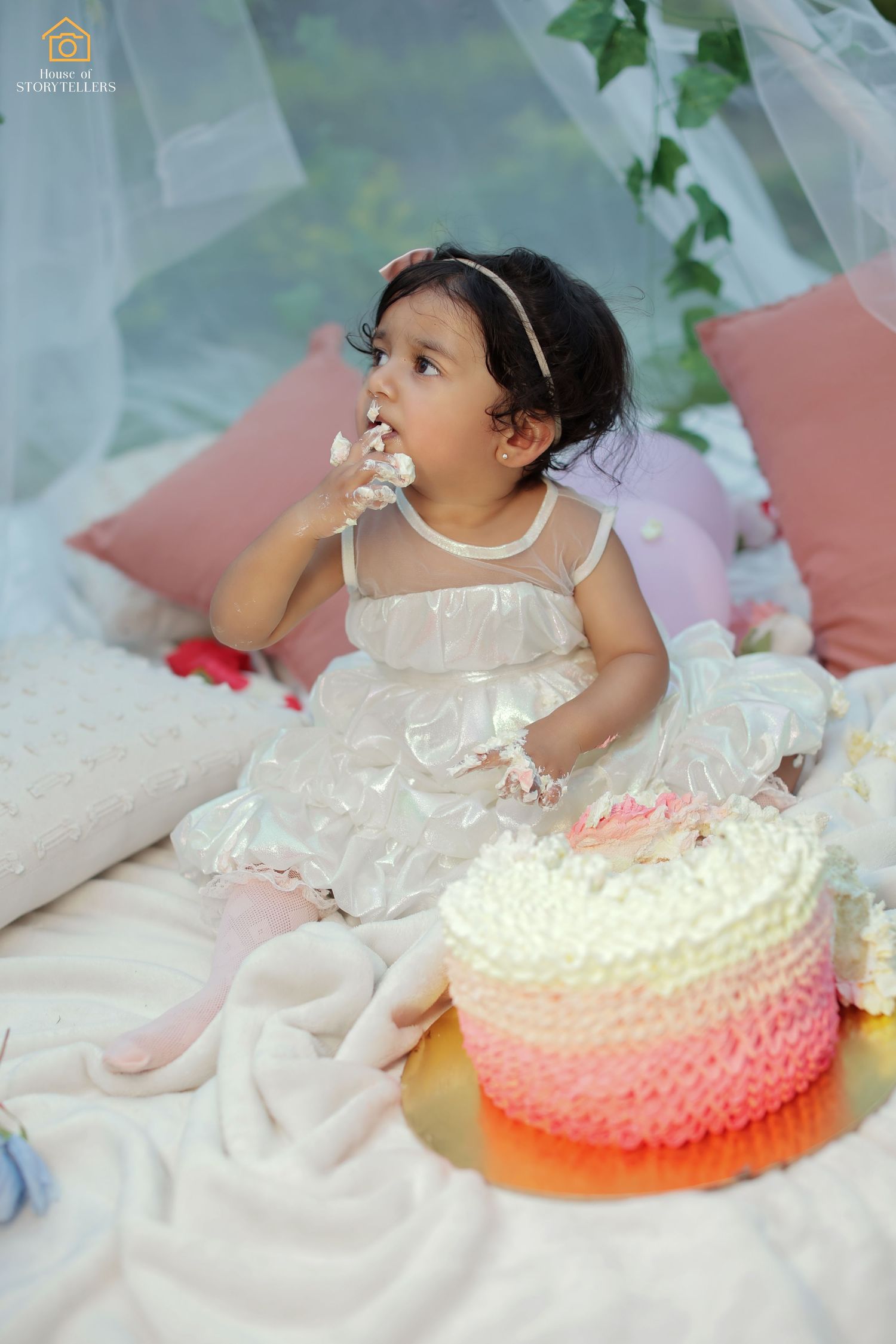 Child in white dress tasting frosting beside an ombre pink celebration cake.