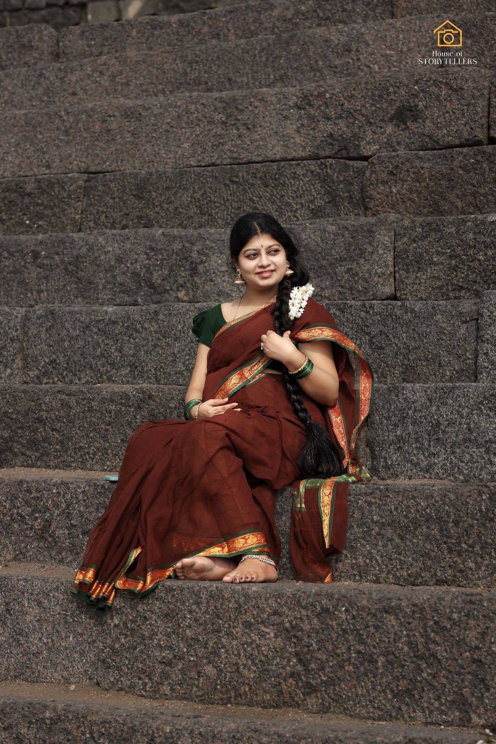 Woman in maroon saree sitting on stone steps against dark wall background.