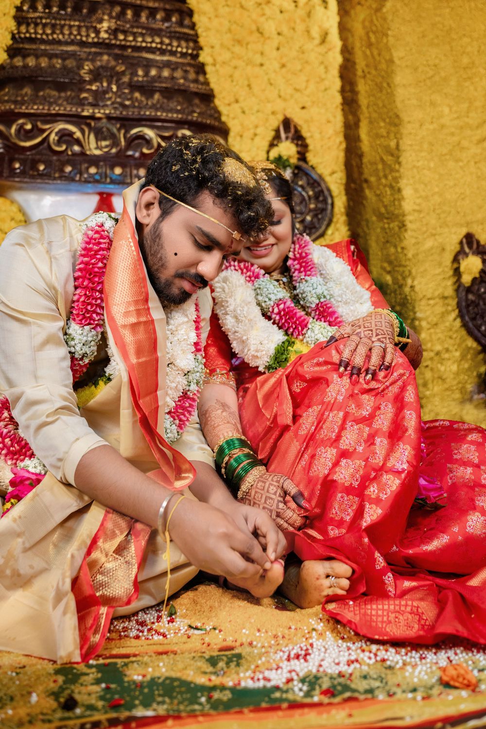Groom placing toe ring on bride during traditional South Indian Hindu wedding ceremony, both wearing floral garlands.