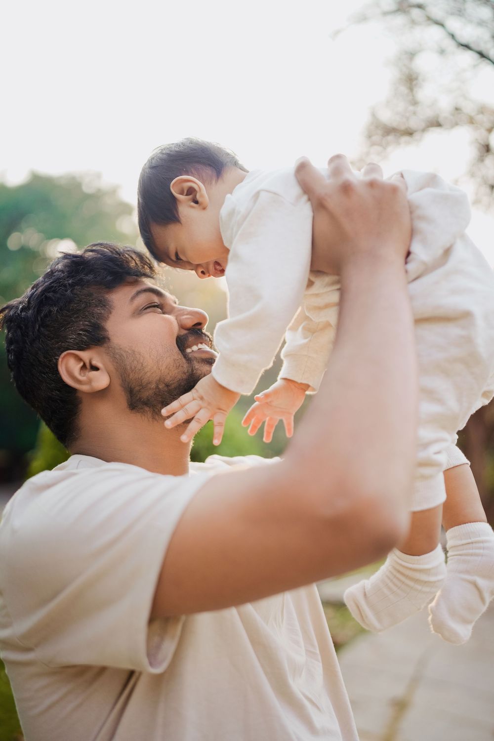 A father joyfully lifts his baby in the air outdoors, both wearing white, sharing a tender moment in a sunlit park.