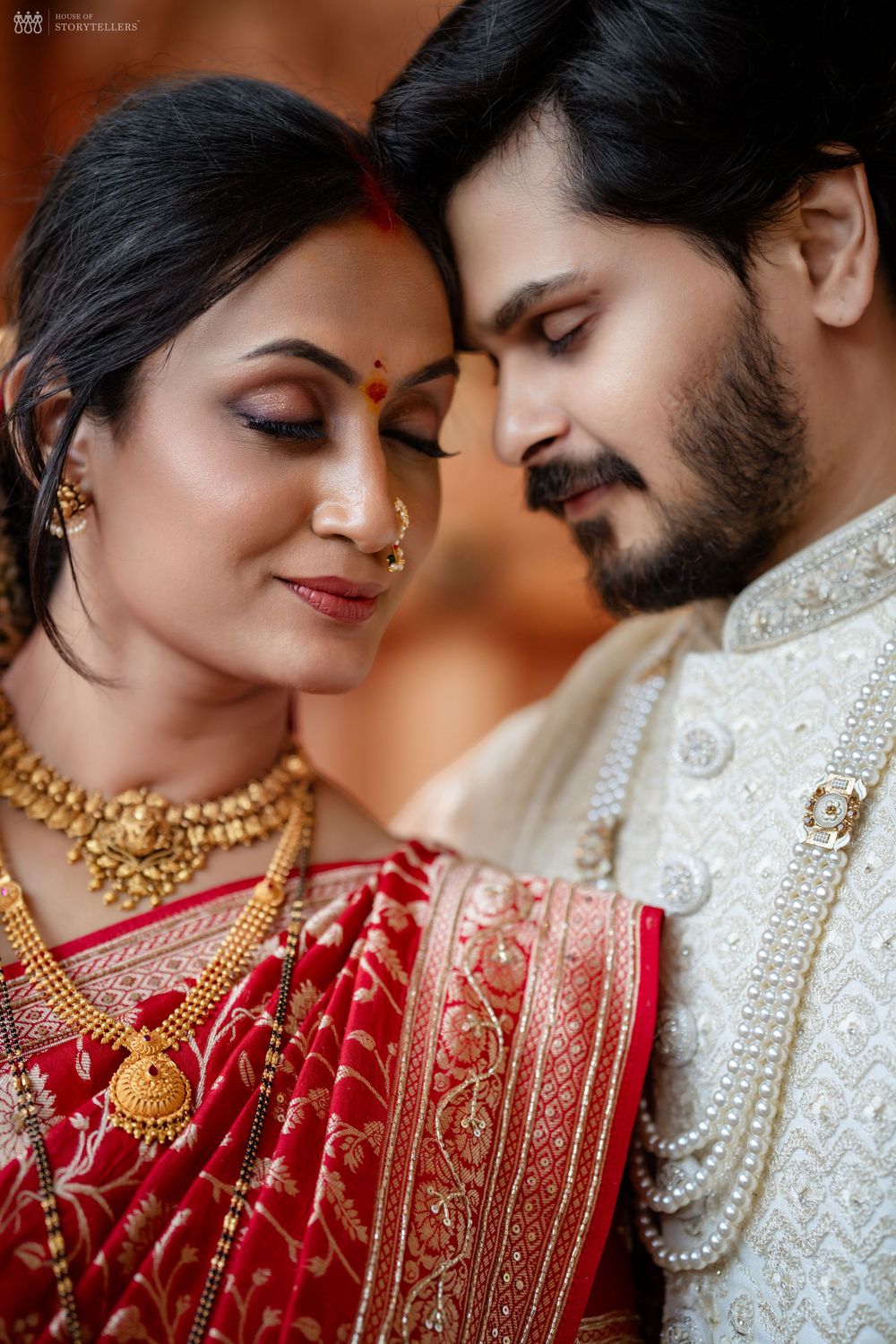 Indian bride in red saree with gold jewelry and groom in white sherwani share an intimate forehead touch moment.