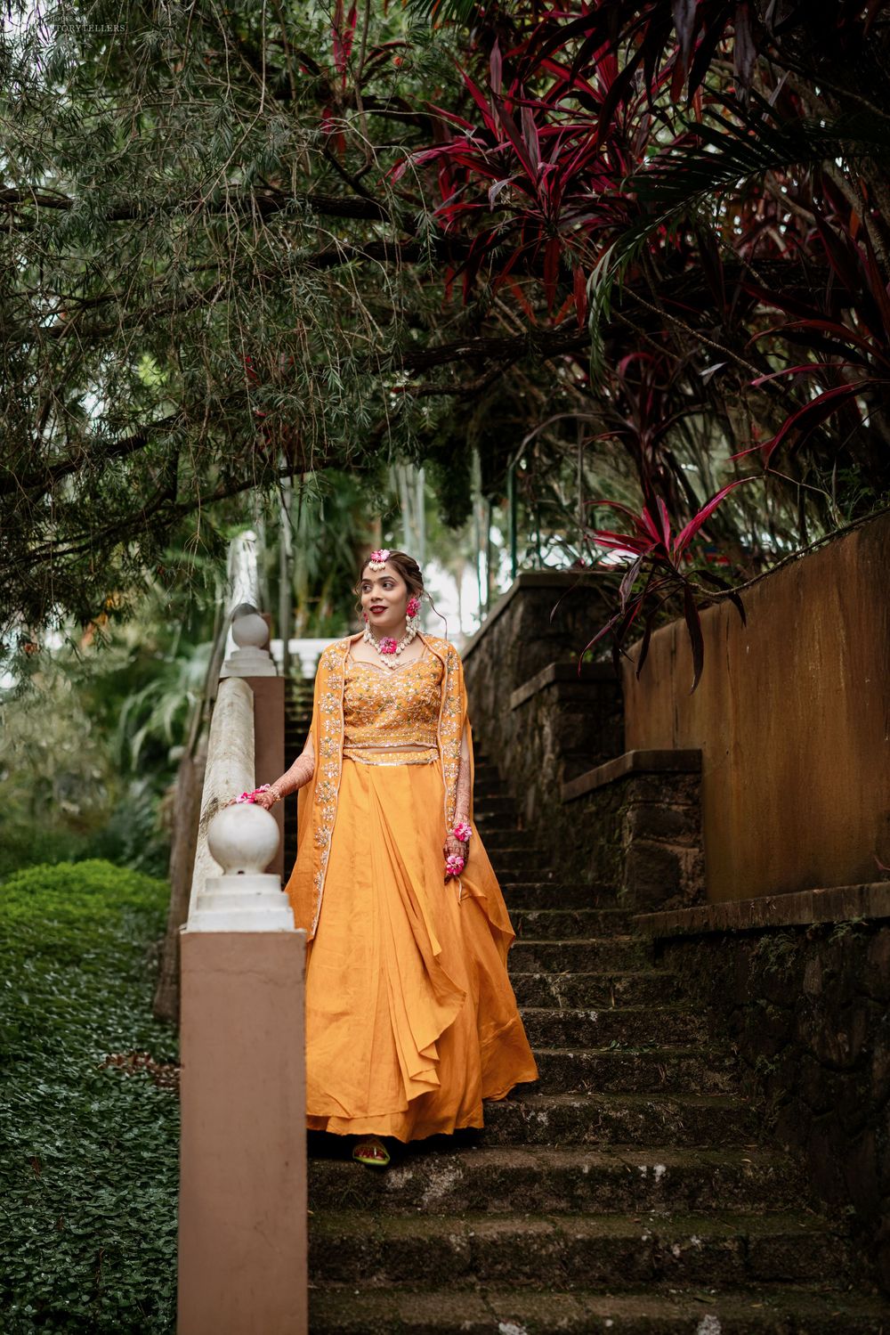 A bride in a vibrant orange lehenga walks down stone steps surrounded by lush greenery and red tropical plants.