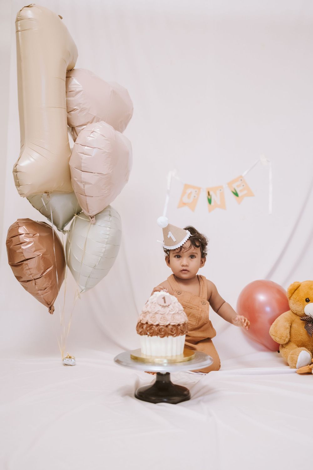A baby in a brown outfit celebrates their first birthday with a smash cake, balloons, and a teddy bear on a white backdrop.