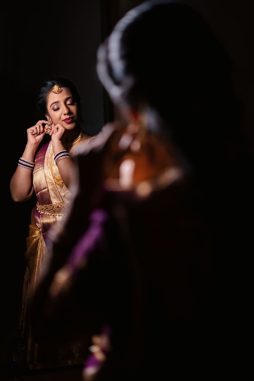 A bride in a golden saree adjusts her earring while her reflection appears in a mirror during bridal preparations.