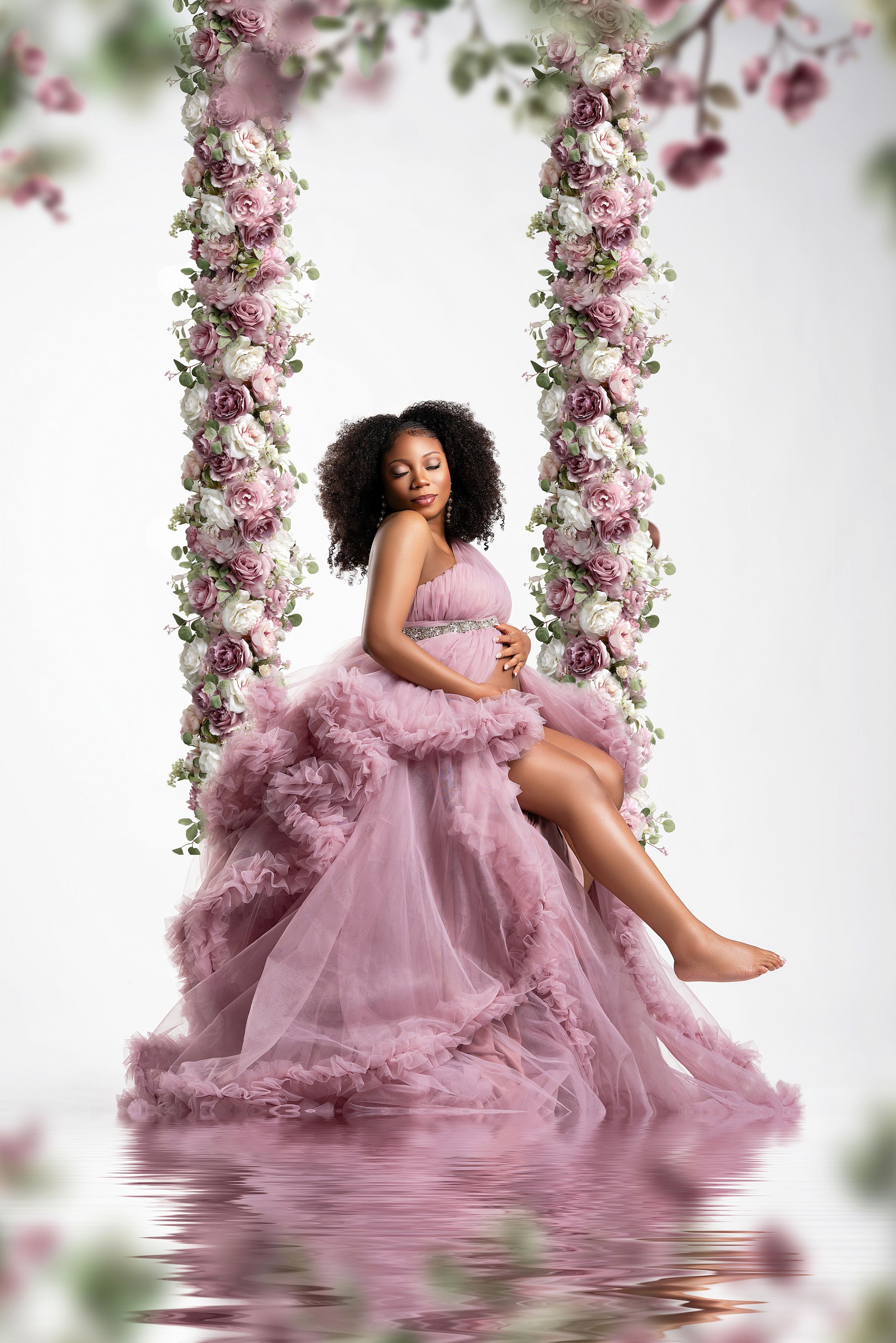 A dreamy maternity portrait with a flowing mauve tulle gown and floral archway reflected in still water.