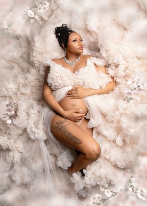 Ethereal maternity photo with flowing white fabric and flowers creates dreamy atmosphere on cloudy background.