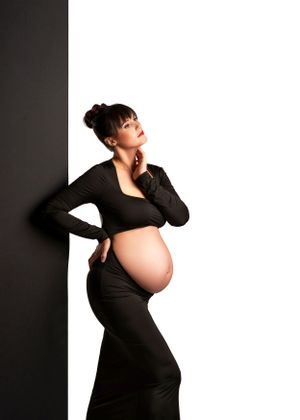 A pregnant woman in a black outfit poses artistically against a white wall in dramatic lighting with a minneapolis maternity photographer