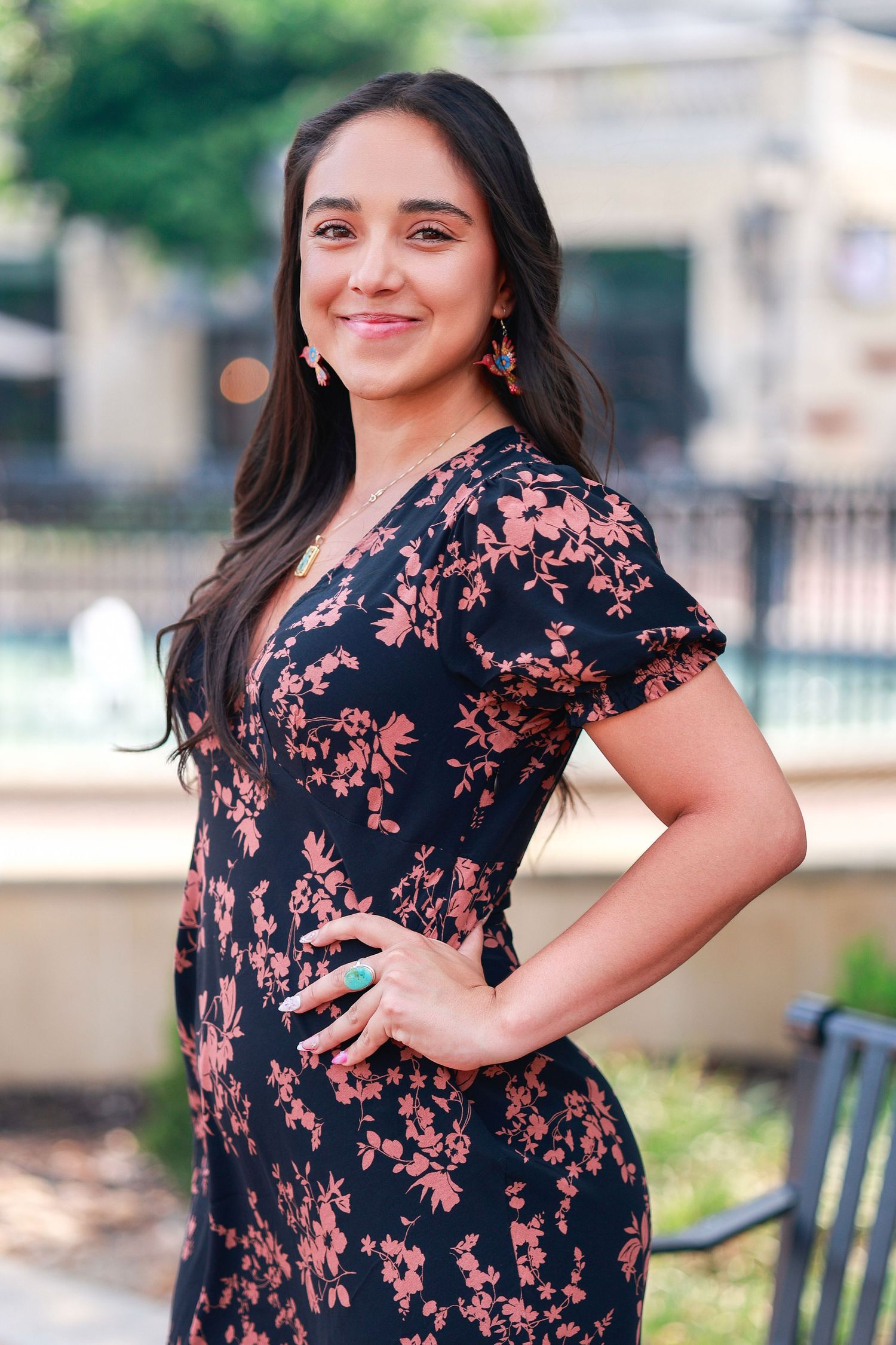 A person stands outdoors smiling confidently while wearing a navy floral dress with pink blooms against a blurred garden setting.