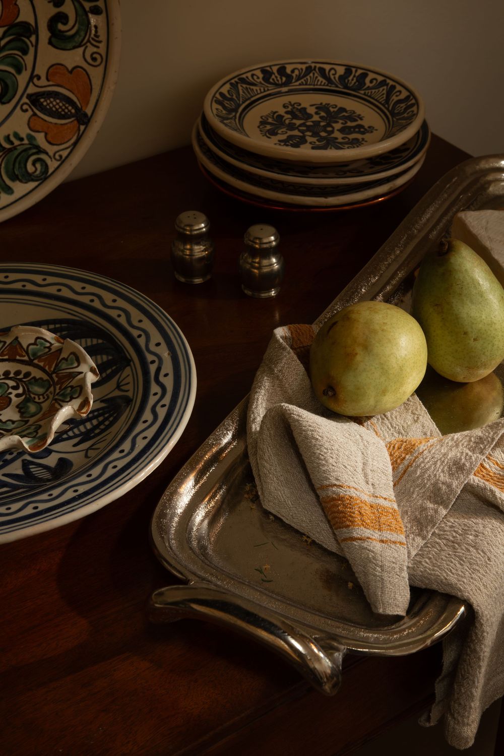Still life with decorative plates, pears, and fabric napkins arranged on a dark wooden surface in moody lighting.