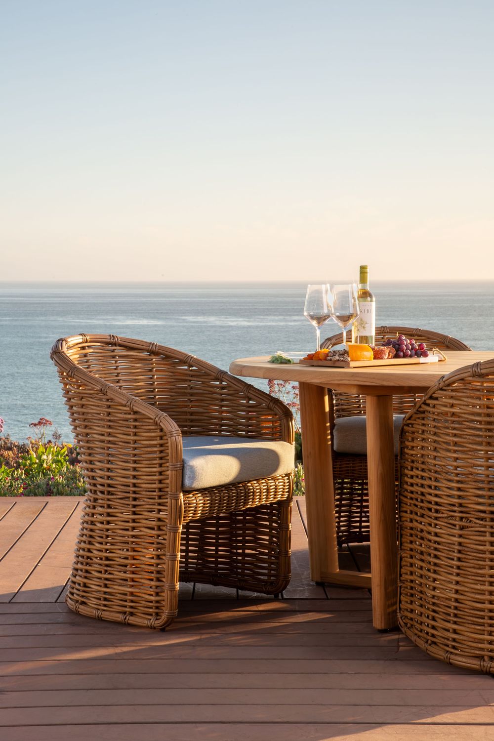 Luxurious outdoor dining setup with wicker chairs and wine glasses overlooking a serene ocean view at sunset.