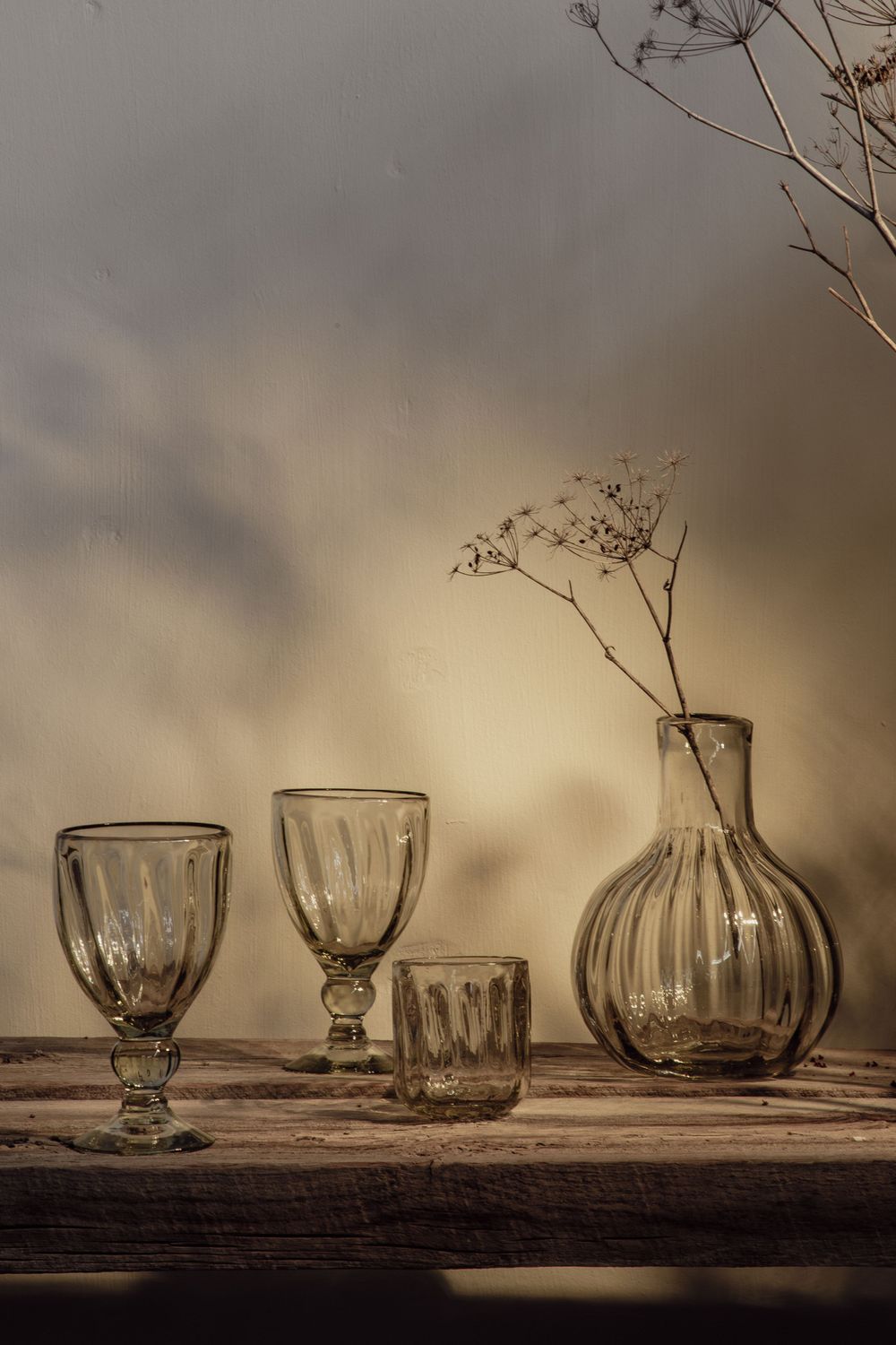 Still life composition with antique glassware including goblets and a ribbed vase with dried flowers on a rustic wooden surface.