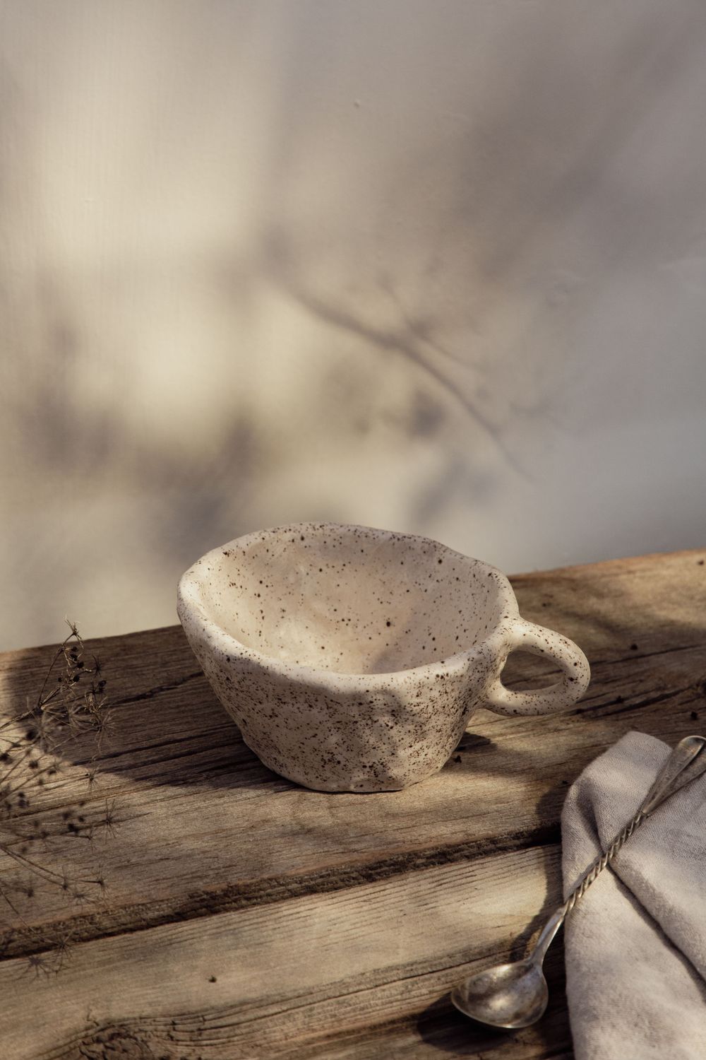 A rustic ceramic teacup sits on weathered wood with a spoon and linen napkin in soft, muted lighting.