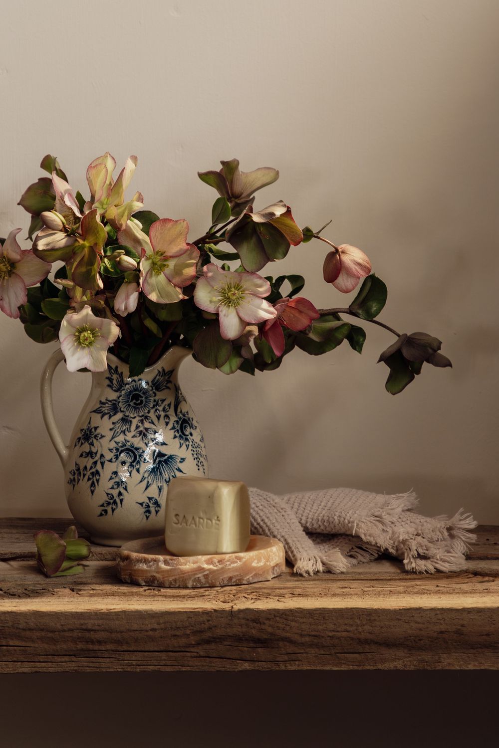 Still life with hellebores and greenery in blue and white ceramic pitcher with soap bar and linen on rustic wooden shelf.