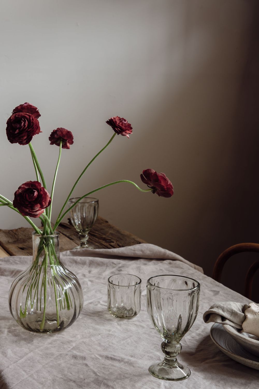 Deep burgundy ranunculus flowers in a clear glass vase with vintage goblets on a white linen tablecloth.