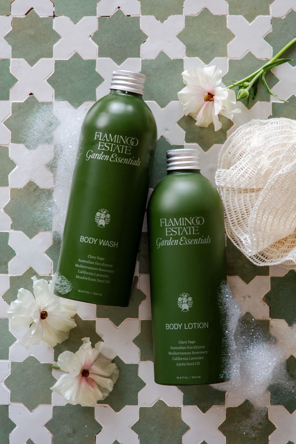 Two green Piantino luxury body lotion bottles displayed on a decorative tile floor with white flowers and a loofah.