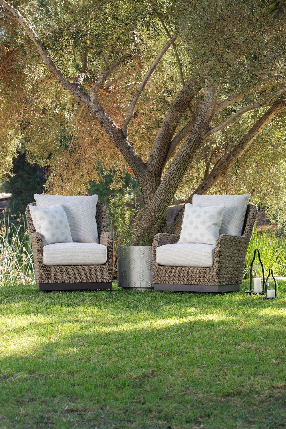 Two wicker patio chairs with plush white cushions sit beneath shady trees on a grassy lawn in an outdoor setting.