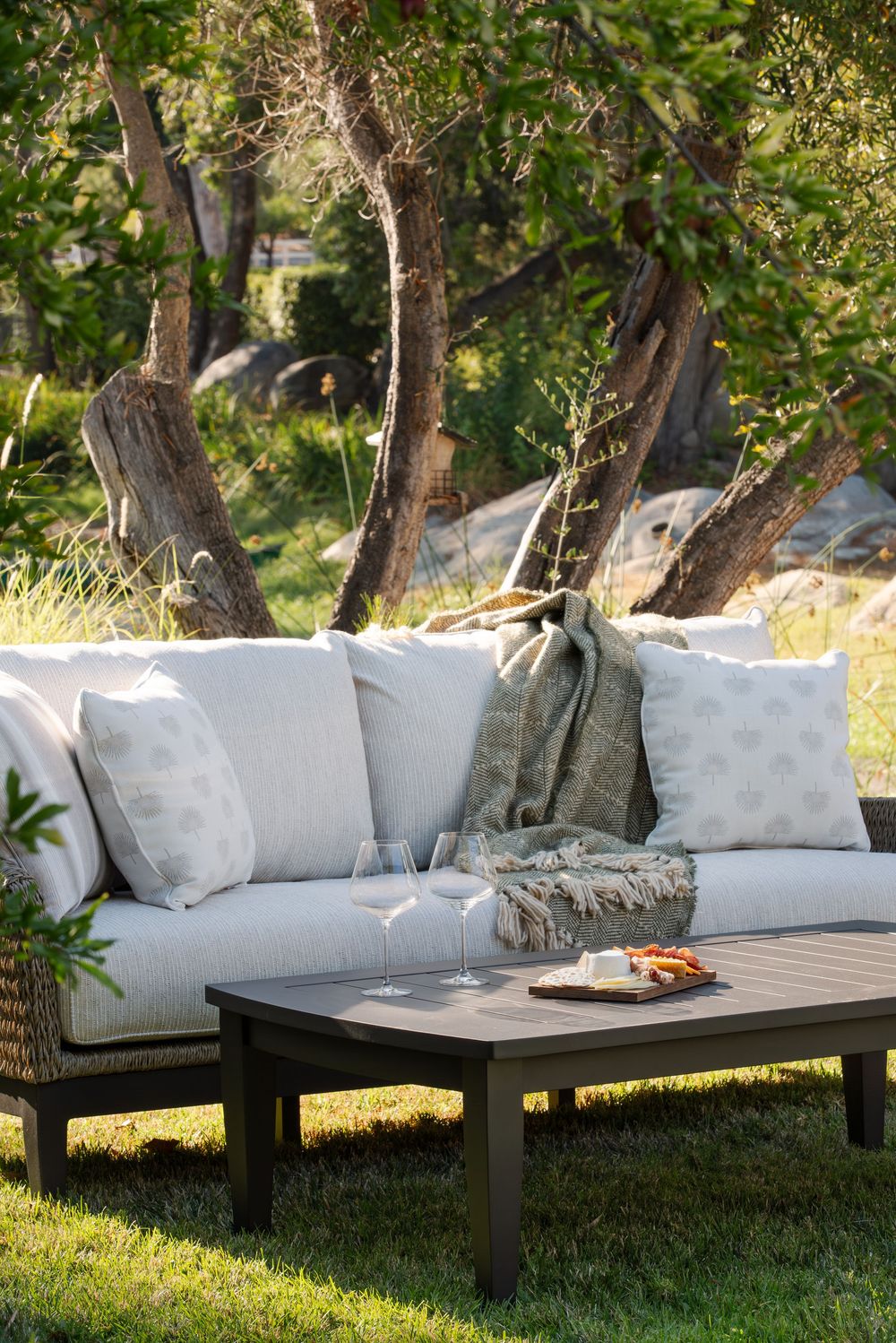 Modern outdoor sectional sofa with white cushions arranged on grass under trees in a tranquil garden setting.
