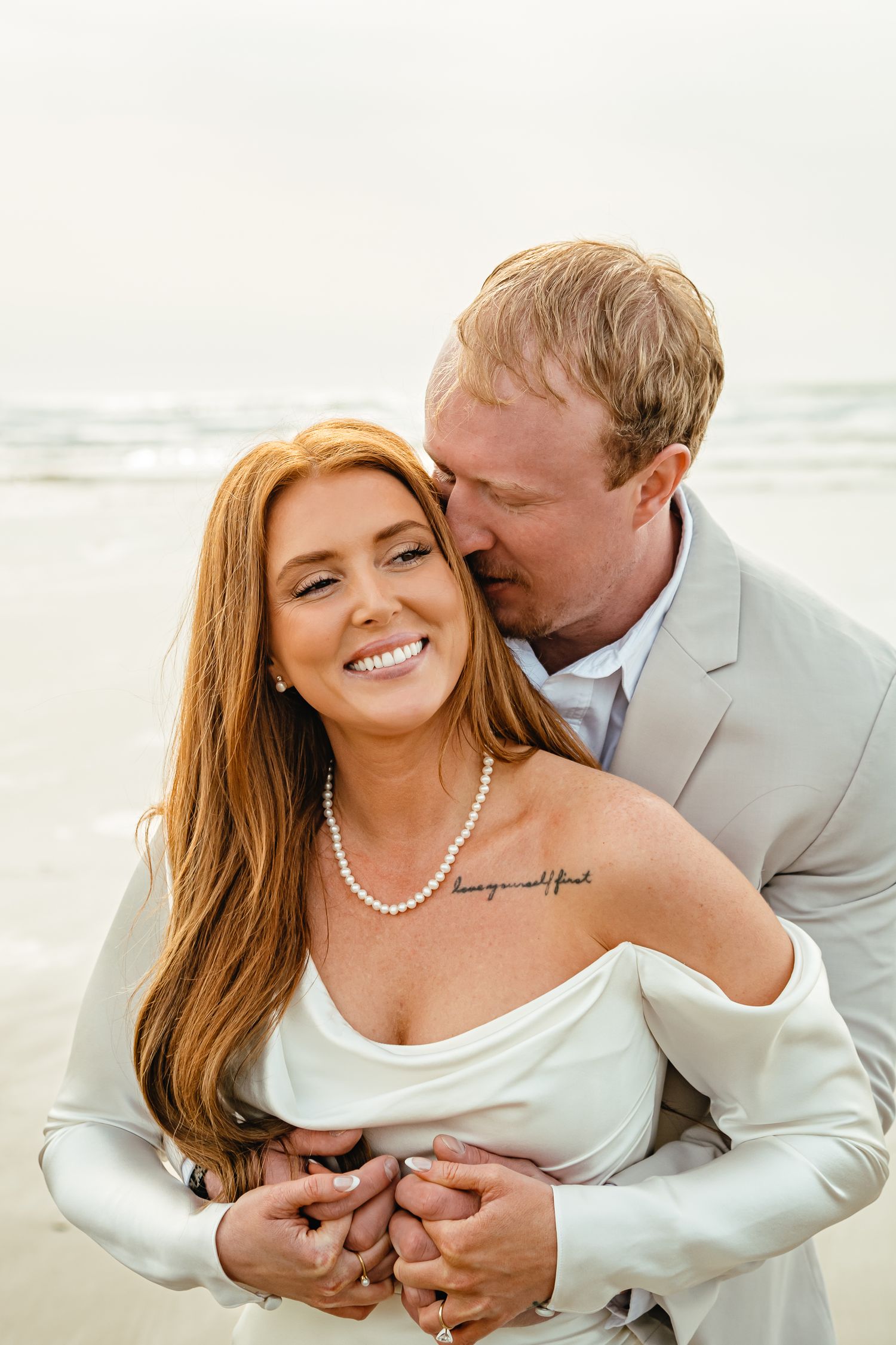 A happy couple embraces on a beach, the bride wearing a white off-shoulder dress and pearl necklace.