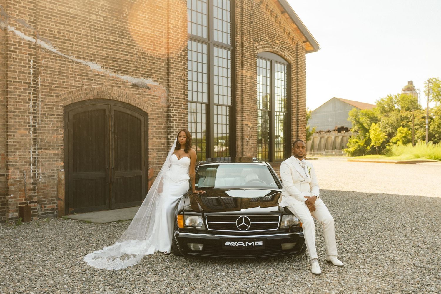 A wedding party poses in front of a rustic brick building with elegant Mercedes-Benz vehicles at sunset. CATSKILLS WEDDING PHOTOS