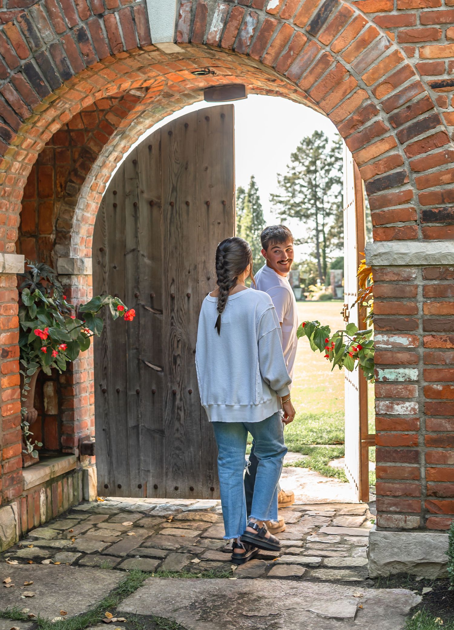Two people stand under a brick archway with a wooden door and flowering vines.