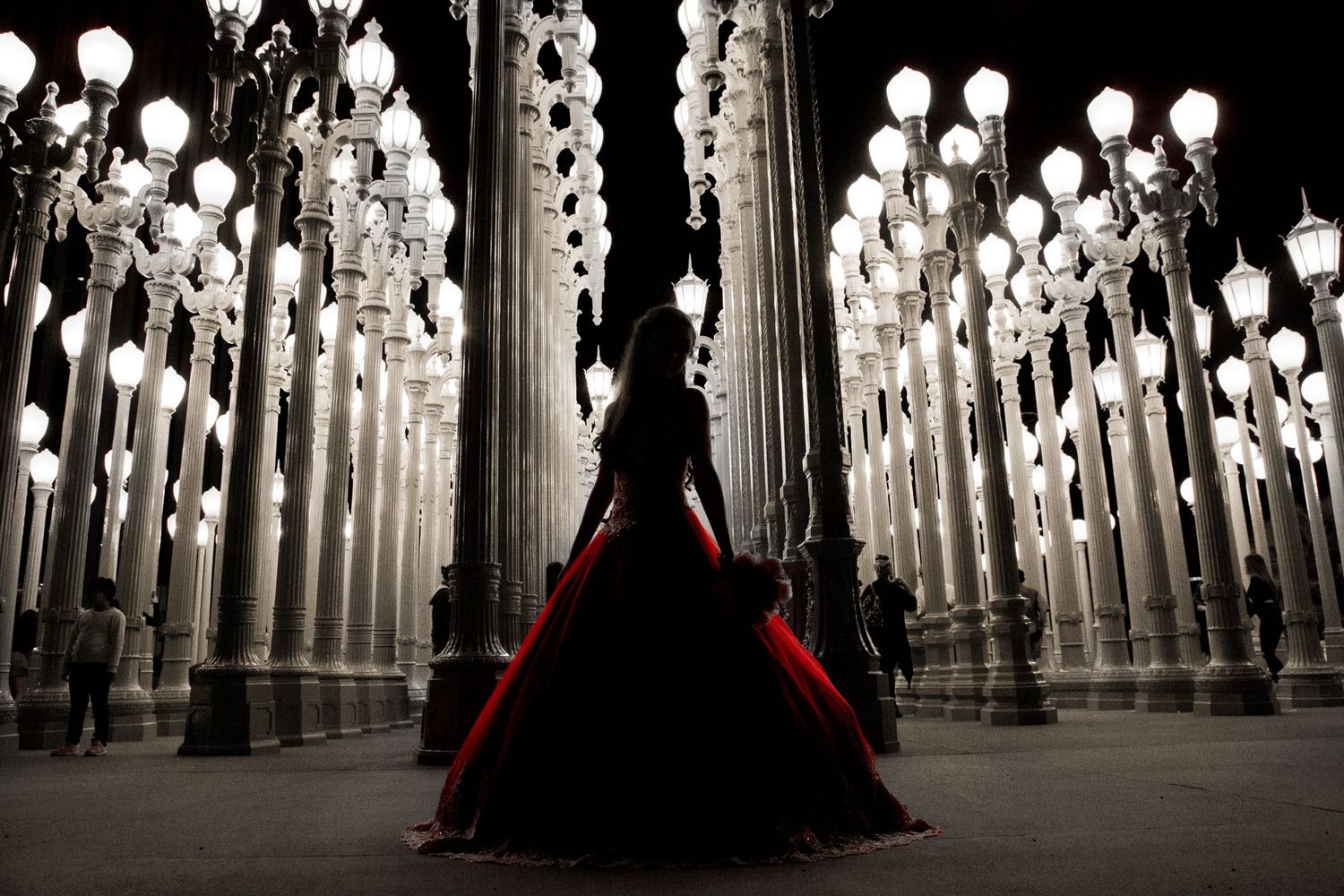 A silhouetted figure in a dramatic black and red gown stands among illuminated white light posts at LACMA's Urban Light installation.