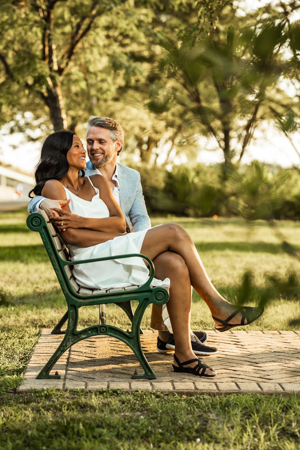 Couple sitting on a green park bench, smiling and embracing in a sunny outdoor setting with lush green trees.