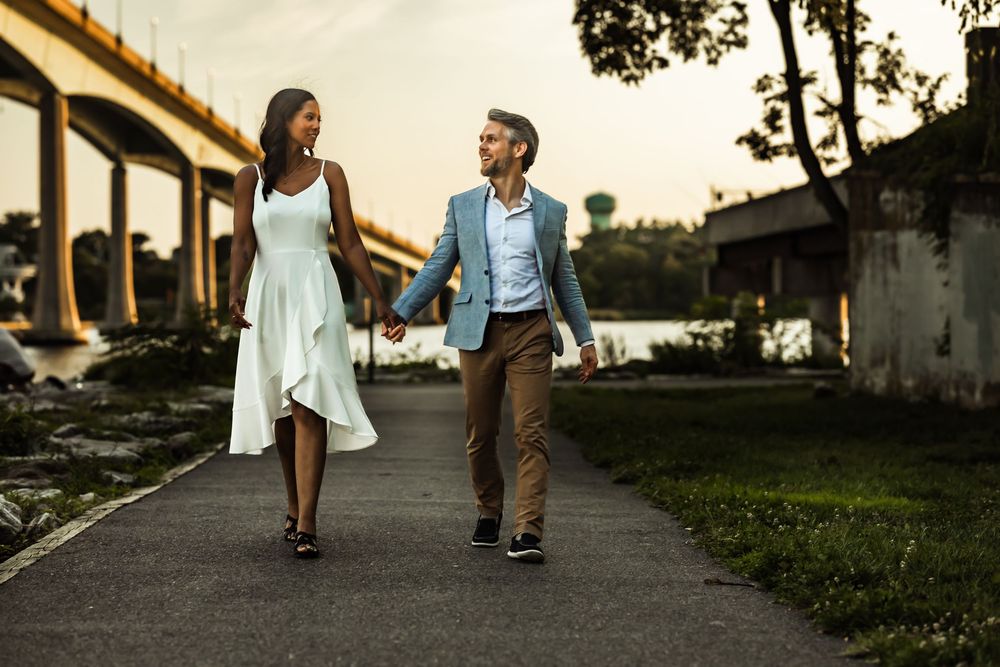 Couple holding hands and walking on a riverside path at sunset near a bridge, woman in white dress, man in blue blazer.