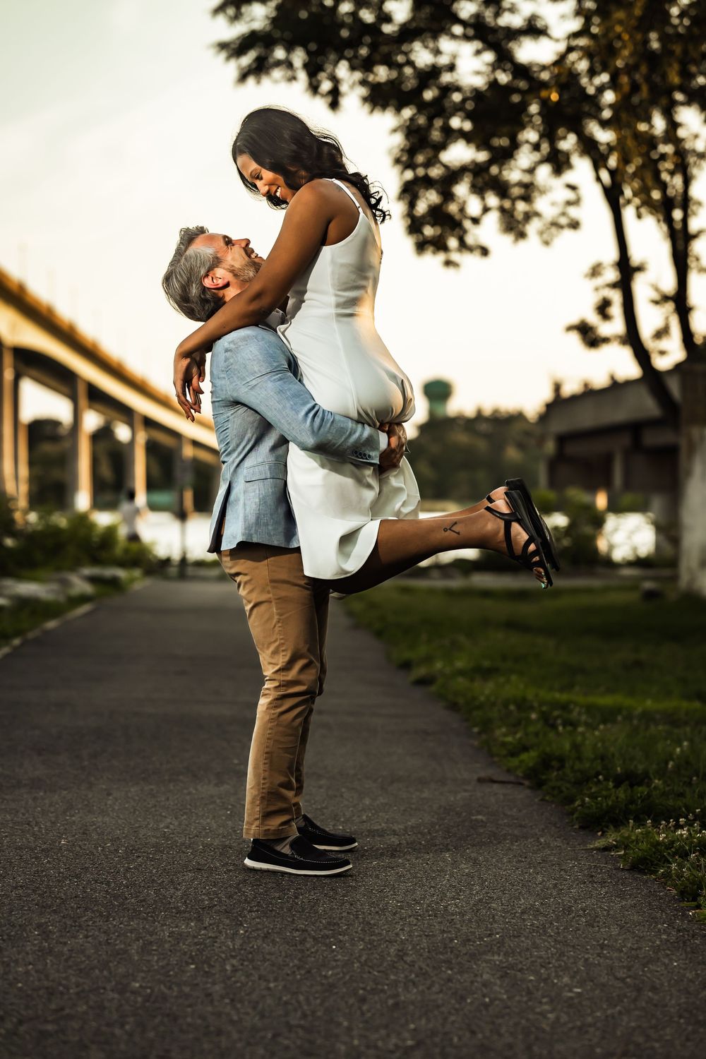 A man lifts a woman in a joyful embrace on a park path at dusk, with a bridge and trees in the background.
