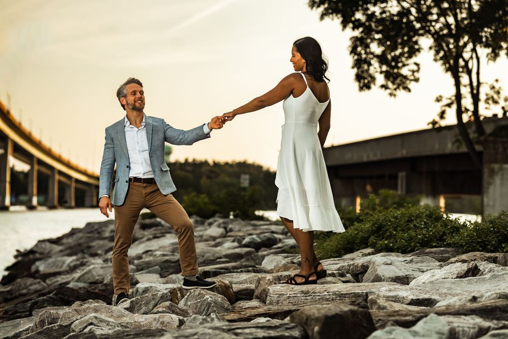 Couple holding hands on rocky waterfront at sunset with bridge in background, man in blazer and woman in white dress.