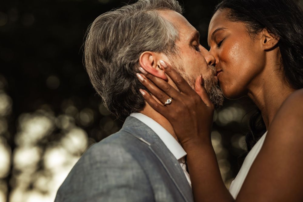 Interracial couple sharing a romantic kiss at their wedding, bride wearing engagement ring.