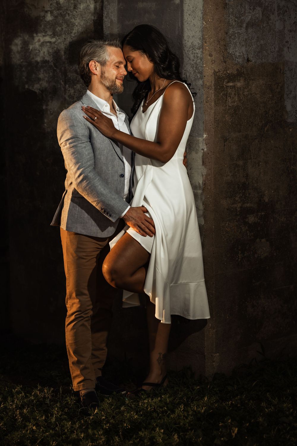 Romantic interracial couple embracing tenderly, man in gray blazer and woman in white dress, against dark textured wall.