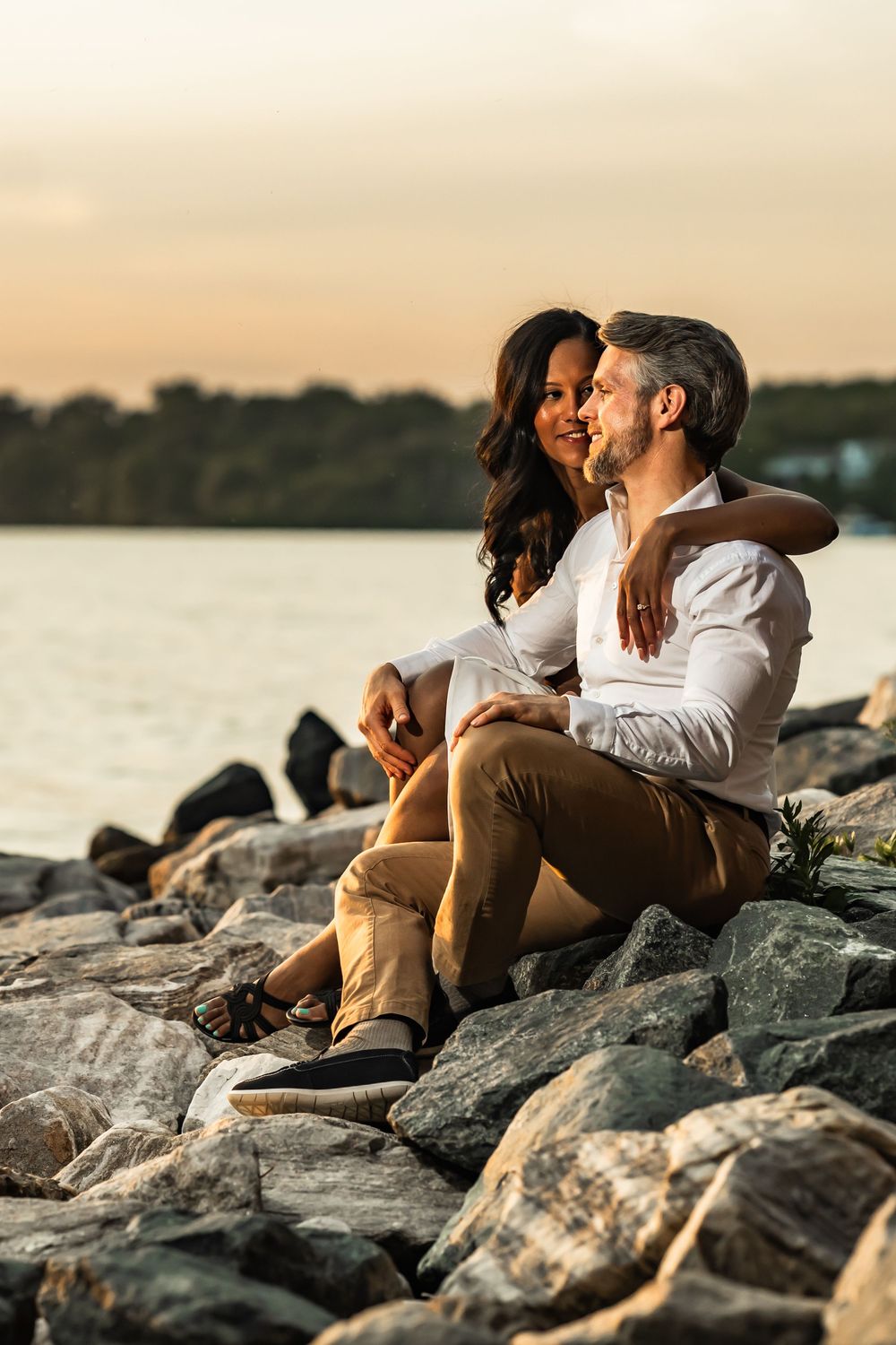 Couple sitting on rocky shoreline at sunset, woman smiling at man while he gazes at the water, trees in background.