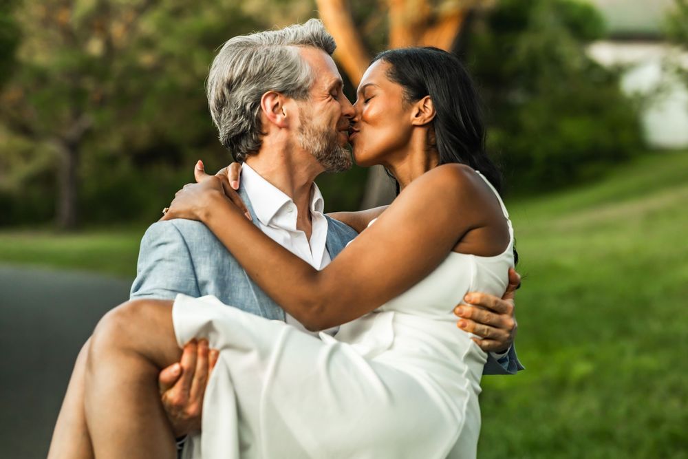 A couple sharing a romantic kiss outdoors, man carrying woman in white dress in a lush green park setting.
