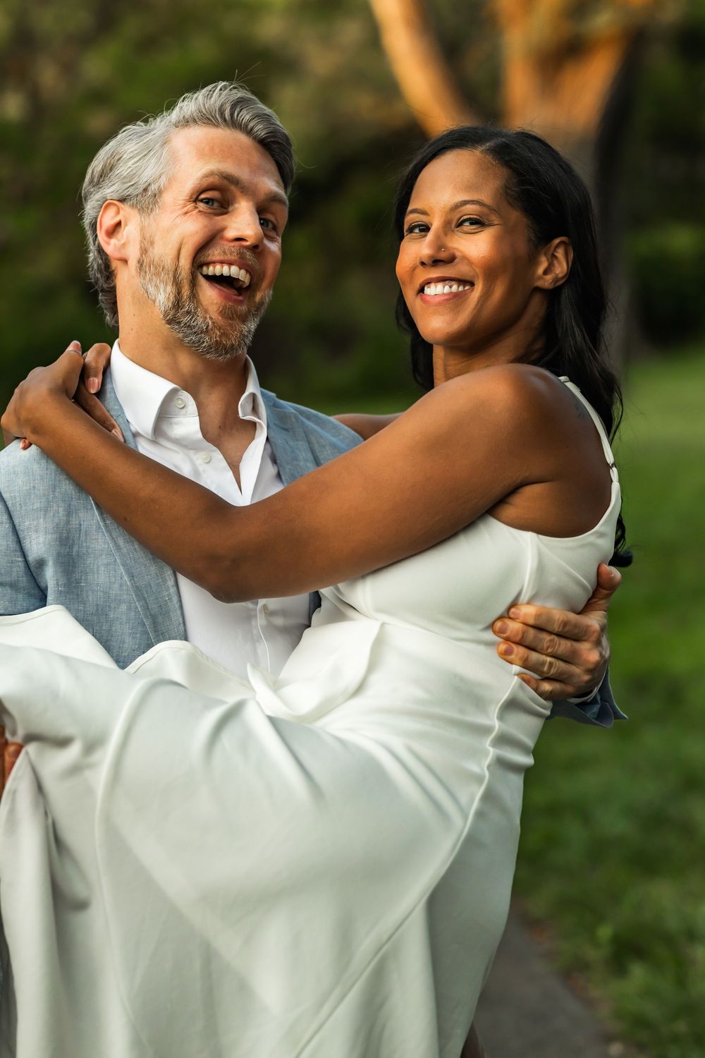 A joyful groom carries his smiling bride in a white dress outdoors, both laughing in a lush green setting.