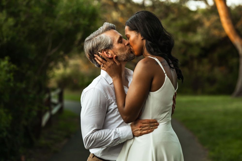 Couple sharing a romantic kiss outdoors, woman in white dress and man in white shirt, surrounded by lush greenery.