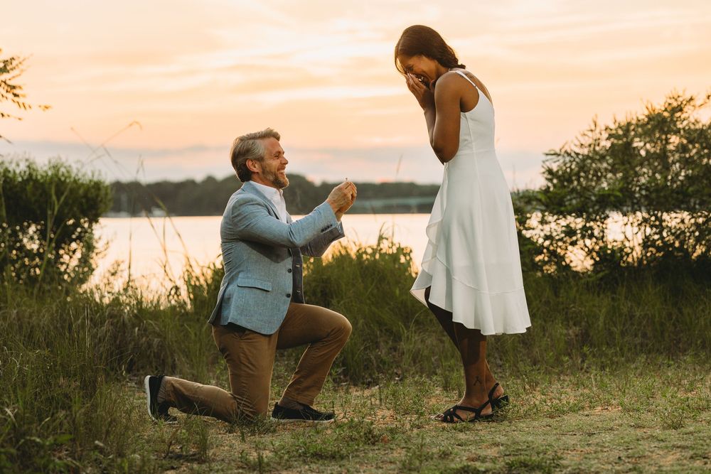 Man kneeling on one knee proposing to woman in white dress at sunset near a lake.
