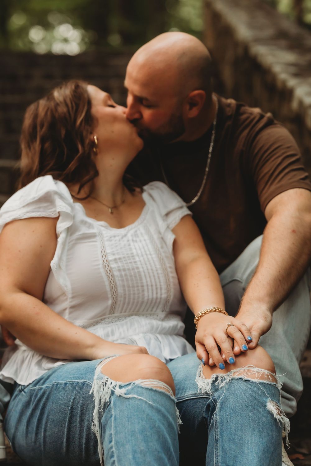 A couple shares a romantic kiss while sitting outdoors, holding hands, wearing casual jeans and tops in a wooded setting.